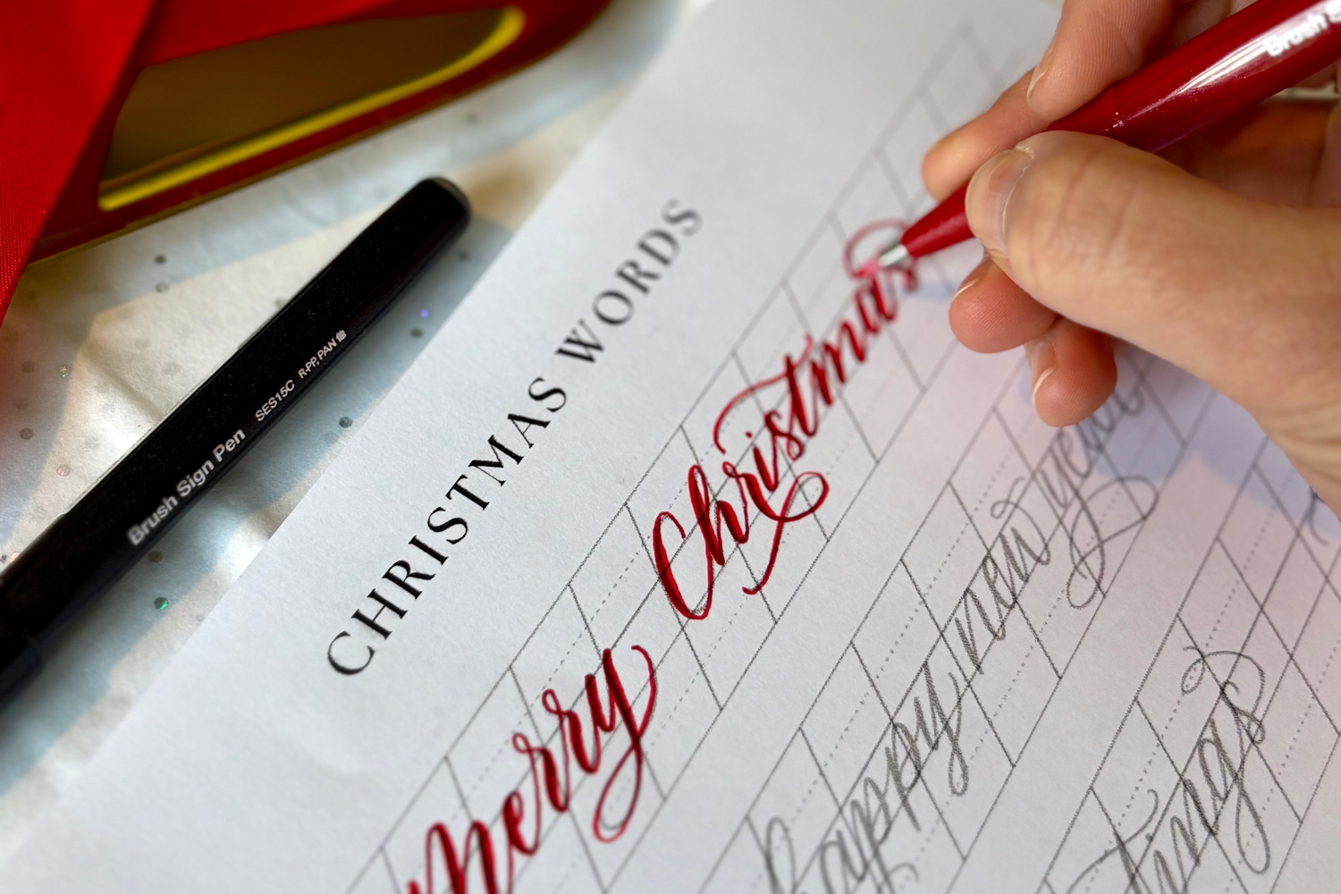 Hand writing 'Merry Christmas' in red ink on a sheet of paper with calligraphy practice. Farnham Surrey Christmas creative art events and workshops with Jodie Rose at Farnham Maltings Museum