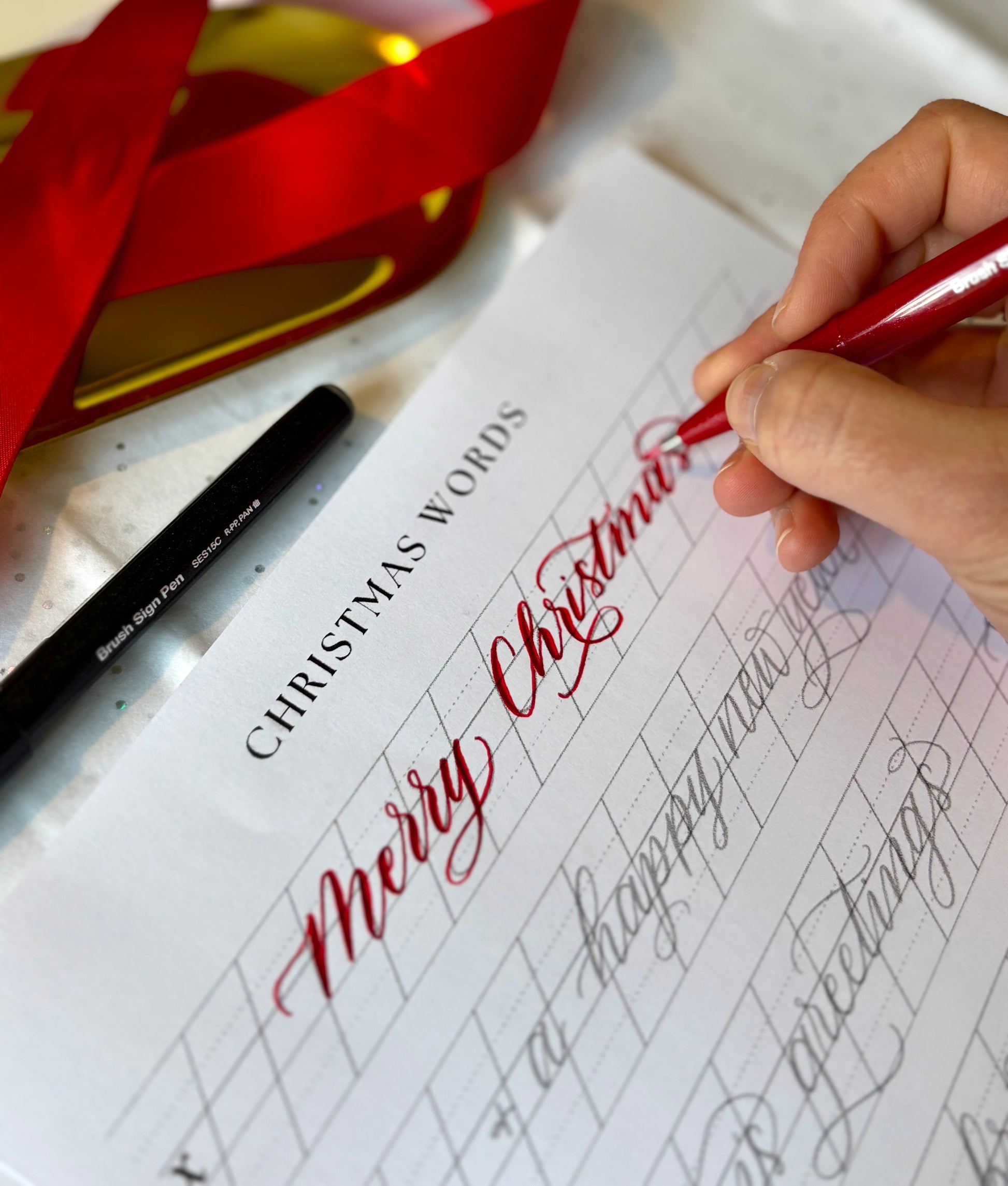 Hand writing 'Merry Christmas' in red ink on a sheet of paper with calligraphy practice. Farnham Surrey Christmas creative art events and workshops 